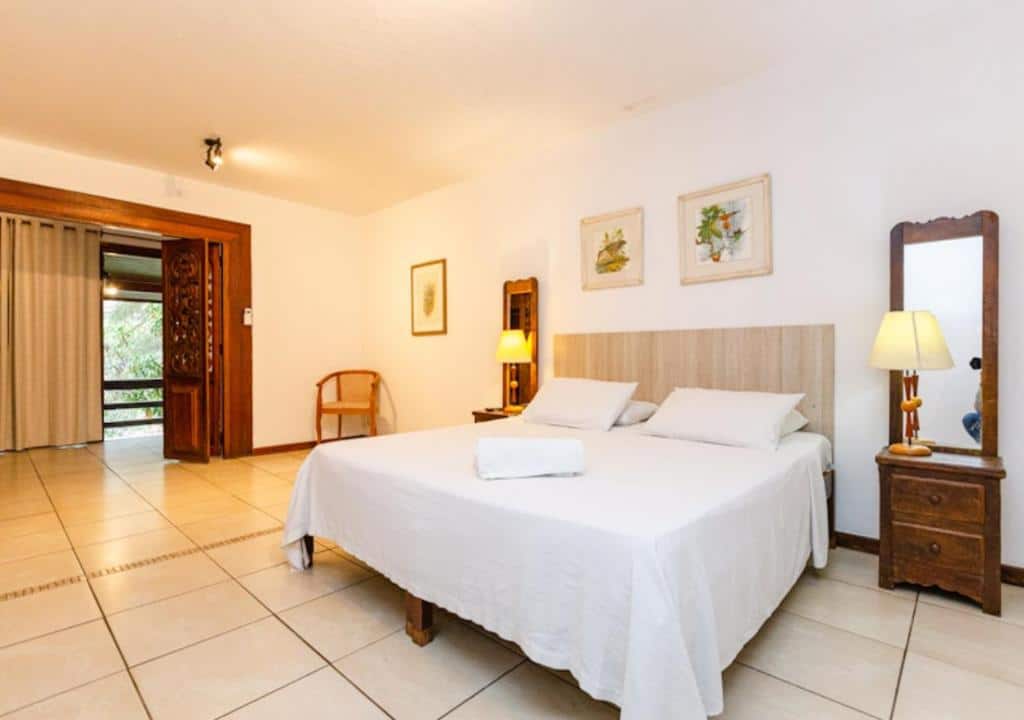Quarto do Hotel Pedra Bonita bem amplo com cama de casal, duas mesinhas de cabeceira com espelho e abajures, além de uma porta que leva até uma varanda, para representar hotéis em Petrópolis