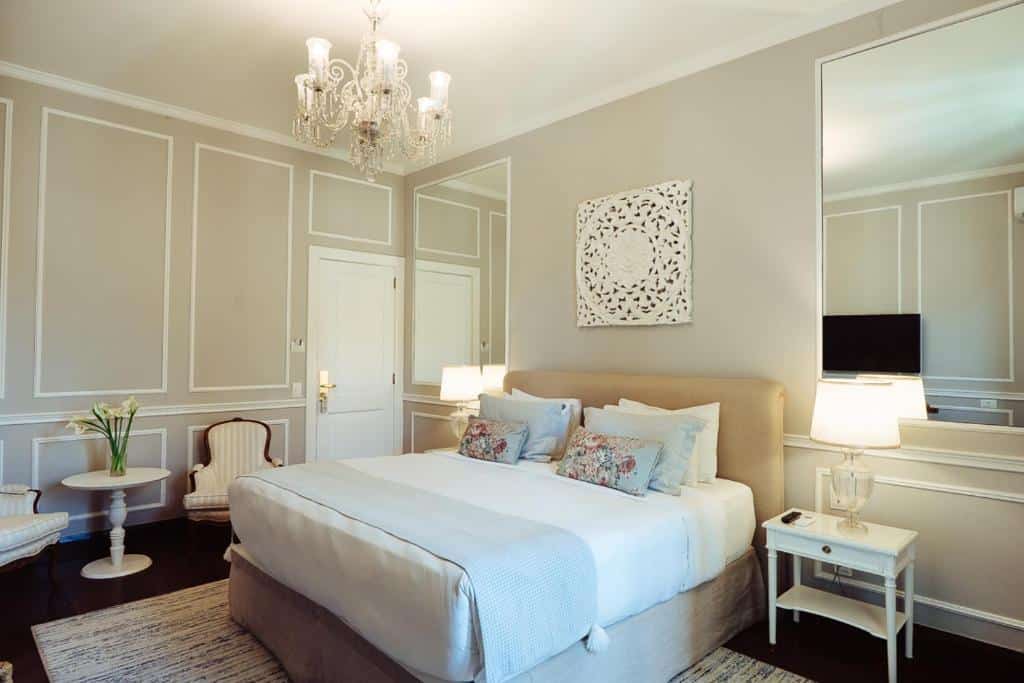 Quarto do Hotel Reggia Catarina com tudo decorado em branco e cinza claro, há um lustre, uma cama de casal, um espelho, duas cabeceiras com abajures, um tapete bege e duas poltronas, para representar hotéis em Petrópolis
