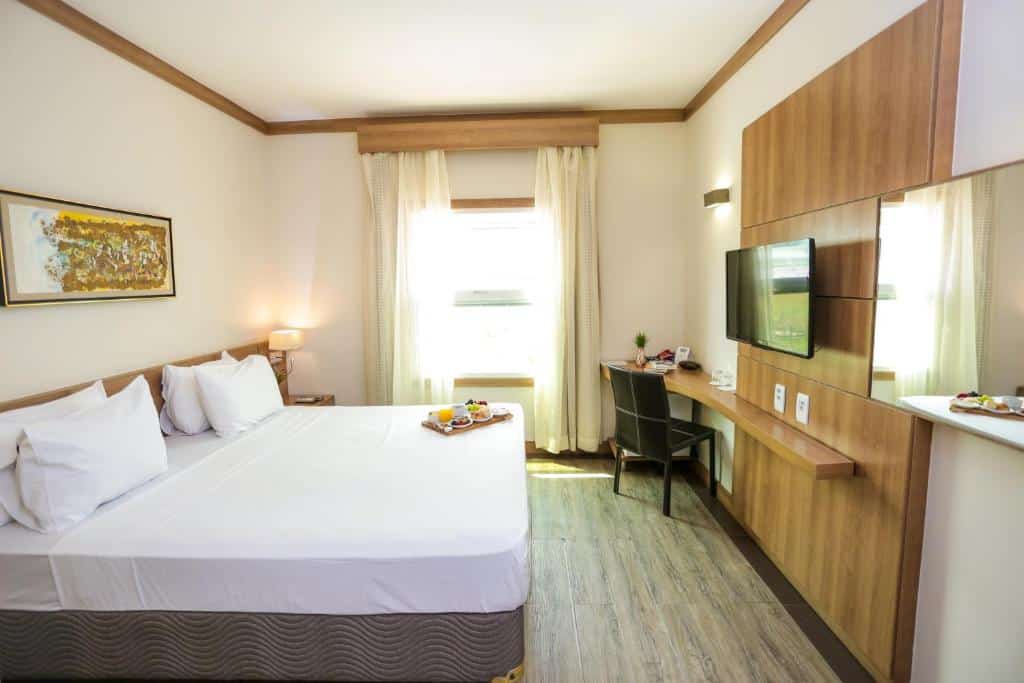 Quarto do Intercity Vinhedo com uma janela com cortinas, um chão de madeira, uma cama de casal, uma bancada com cadeira e uma televisão, para representar hotéis perto do Hopi Hari