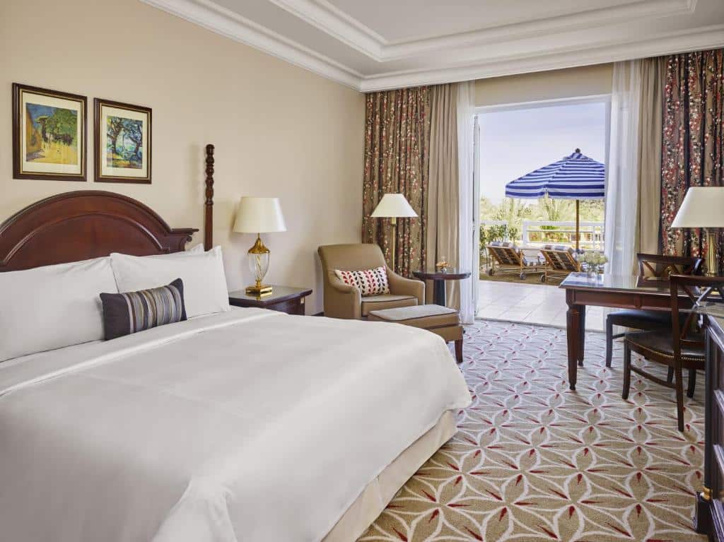 Quarto do JW Marriott Hotel com cama de casal, uma cômoda de madeira do lado esquerdo da cama com luminária, poltrona bege com mesa redonda ao lado das portas que dá acesso a sacada com cortinas branca e marrom.