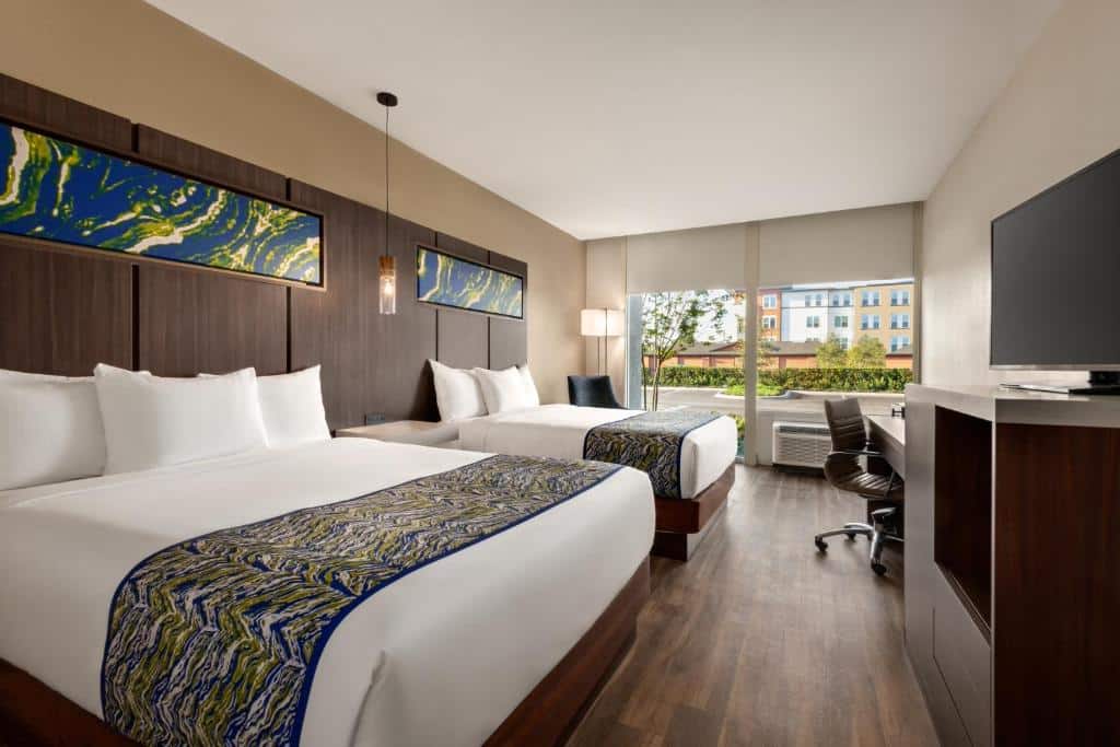 Quarto do La Quinta Inn & Suites by Wyndham Orlando com uma cama de casal, uma de solteiro, chão de madeira, uma janela ampla com cortinas, uma mesa de escritório com cadeira giratória e um móvel de madeira com uma televisão, para representar hotéis perto do SeaWrold