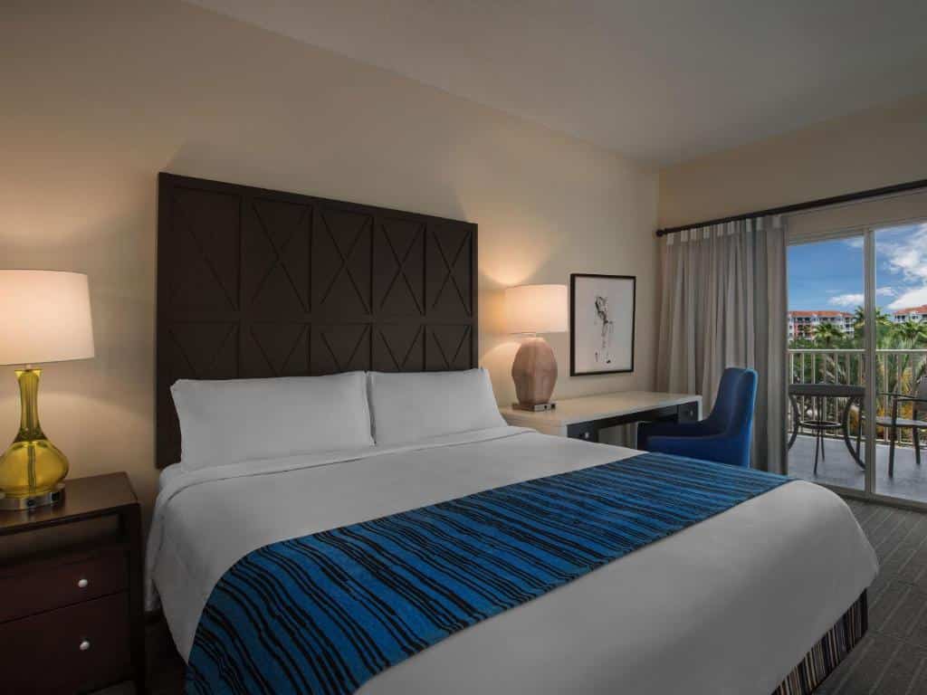 Quarto do Marriott's Grande Vista com uma cama de casal, uma sacada com mesinha e duas cadeiras, chão de madeira, mesa de escritório com cadeira e dois abajures, para representar hotéis perto do SeaWorld