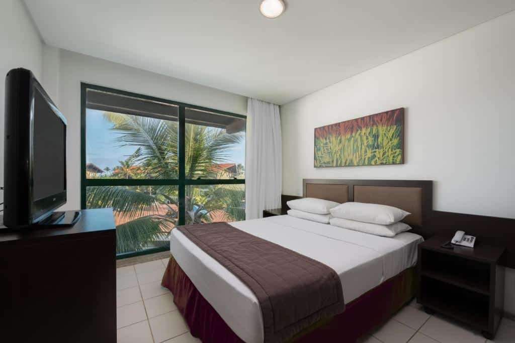 Quarto no Marulhos Suítes Resort by MAI com cama de casal, uma janela ampla com cortinas, e uma móvel cm uma televisão de frente para a cama