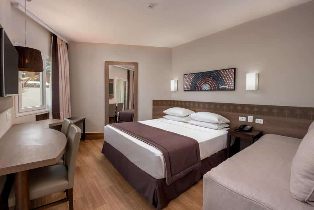 Quarto do Marupiara Resort com uma cama de casal, chão de madeira, uma cabeceira com duas luminárias, uma espelho de corpo inteiro e uma televisão presa na parede de frente para a cama, para representar resorts em Muro Alto