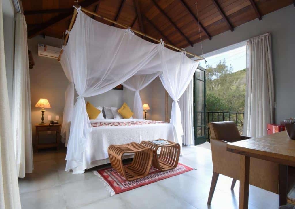 Quarto da Pedra da Laguna Boutique Hotel SPA com uma sacada, uma cama ampla com bangalô de tecidos brancos, duas mesinhas de cabeceira com abajures, dois bancos rústicos de madeira, para representar resorts em Búzios