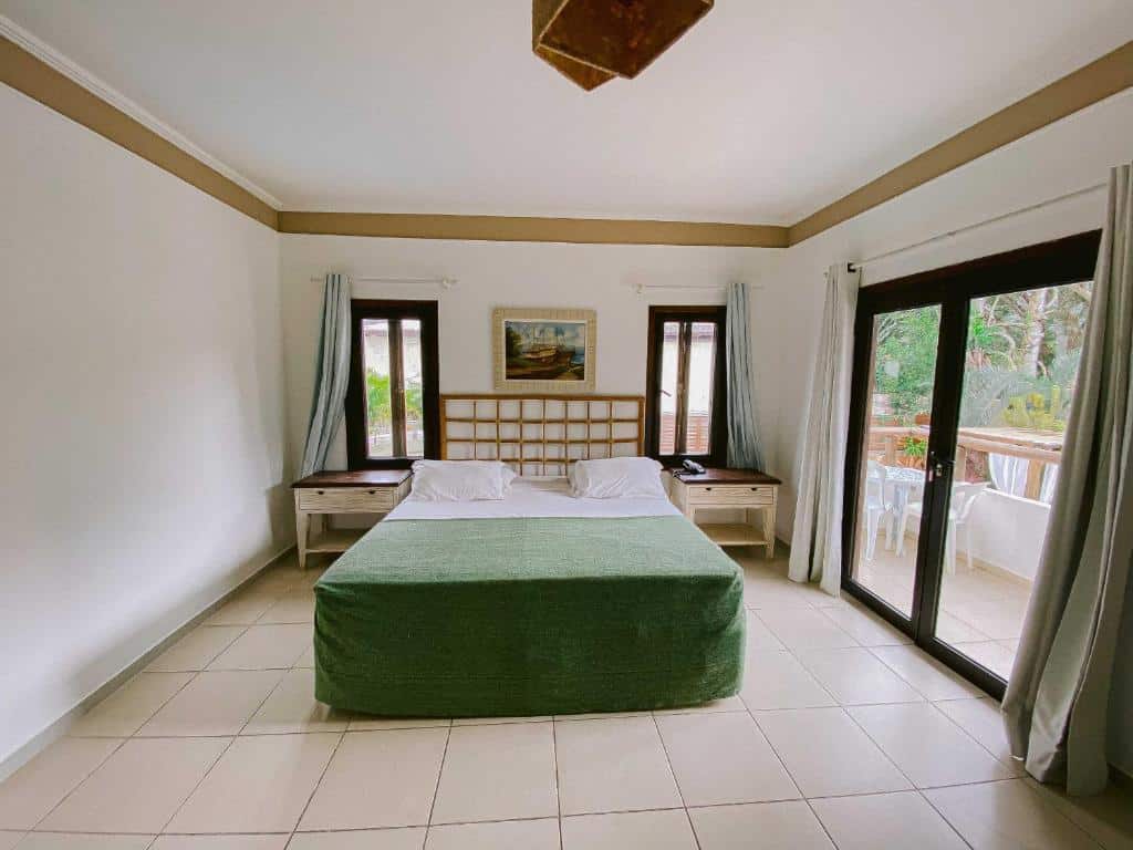 Quarto da Pousada Porto Mare com cama de casal no centro do quarto, duas cômodas de madeira do lado da cama, do lado direito portas de vidro com cortinas verde claro.