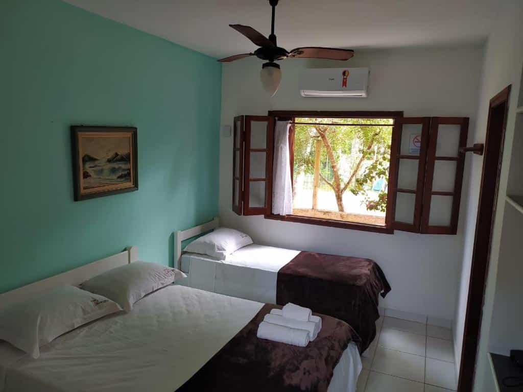 Quarto da Azul Banana com cama de casal do lado direito, do lado esquerdo cama de solteiro e janela de madeira aberta. No centro do quarto ventilador de teto.