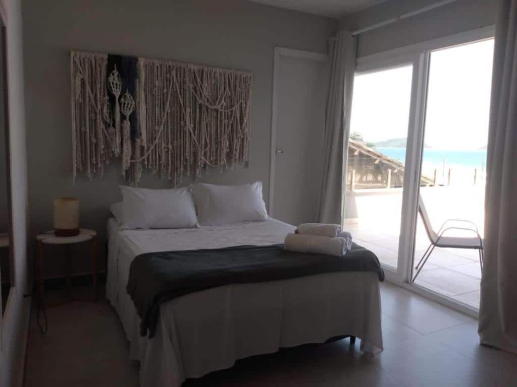Quarto da Casatua com cama de casal, mesa do lado esquerdo com luminária e do lado direito portas de vidro com cortinas brancas que dá acesso a varanda térreo com acesso a praia.