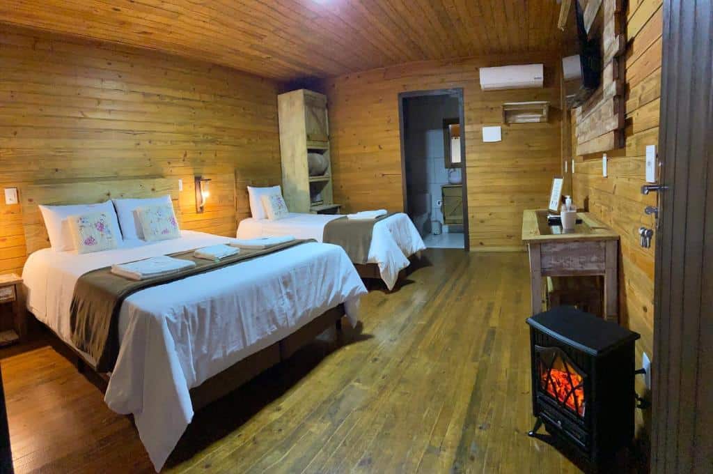 interior do chalé na Pousada Costaneira com uma cama de casal, uma cama de solteiro, uma lareira móvel pequena à direita e uma porta com vista para o banheiro na parede dos fundos do quarto