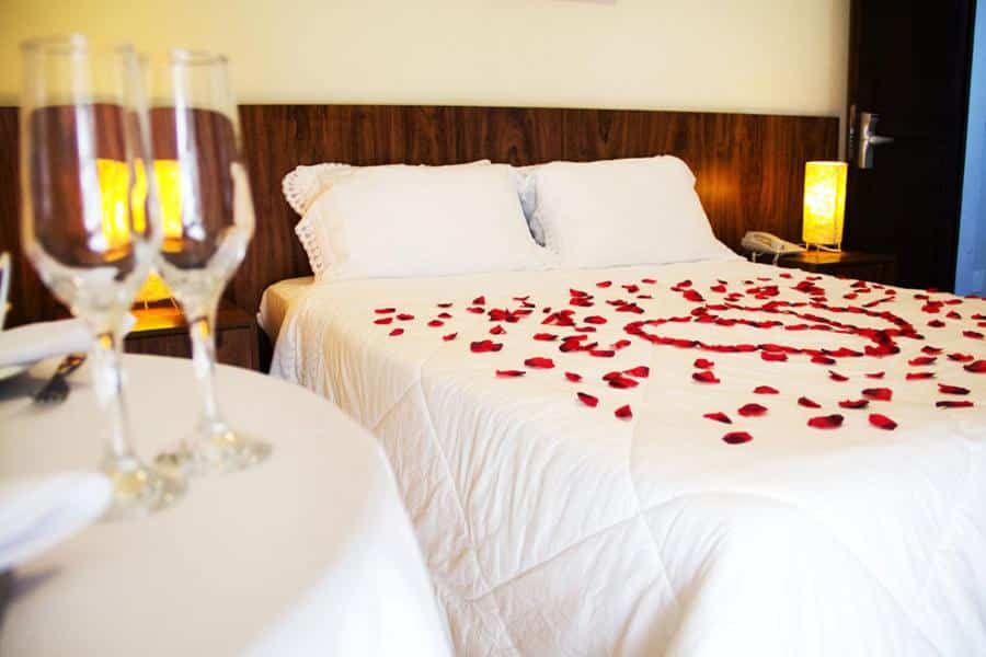 Quarto da Pousada Iguatiba com uma cama de casal cheia de pétalas de flores por cima, além de duas mesinhas de cabeceira com abajures e uma mesa para duas pessoas com taças em cima