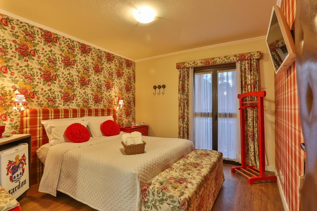 Quarto da Pousada Jardon com tudo decorado com estampas floridas, uma varanda com cortinas, uma cama de casal, um cabideiro, uma televisão, um frigobar e um pequeno sofá