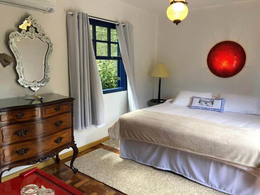 Quarto na Pousada Vila Brasil com uma cama de casal, uma janela com cortinas, uma cômoda estilo antigo com um espelho, chão de taco com tapetes beges, para representar chalés em Petrópolis