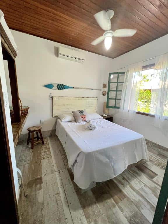 Quarto da Pousada Vila Campeche com uma cama de casal, uma janela ampla com cortinas, chão de madeira clara, ventilador do teto e também ar-condicionado de teto, para representar pousadas na Praia do Campeche
