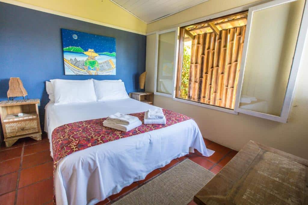Quarto da Pousada Vila Tamarindo Eco Lodge com uma cama de casal com travesseiros e toalhas sob ela, uma janela ampla, uma mesa de madeira envelhecida e dois móveis de cabeceira nas laterais da cama com abajures rústicos, para representar pousadas na Praia do Campeche