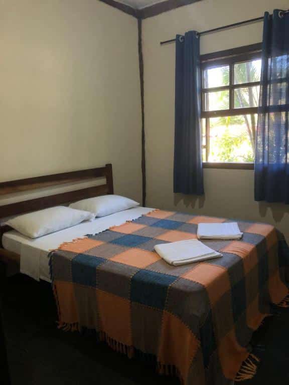 Quarto da Vila Cambury com cama de casal ao centro do quarto e uma janela com cortinas azuis no lado esquerdo.