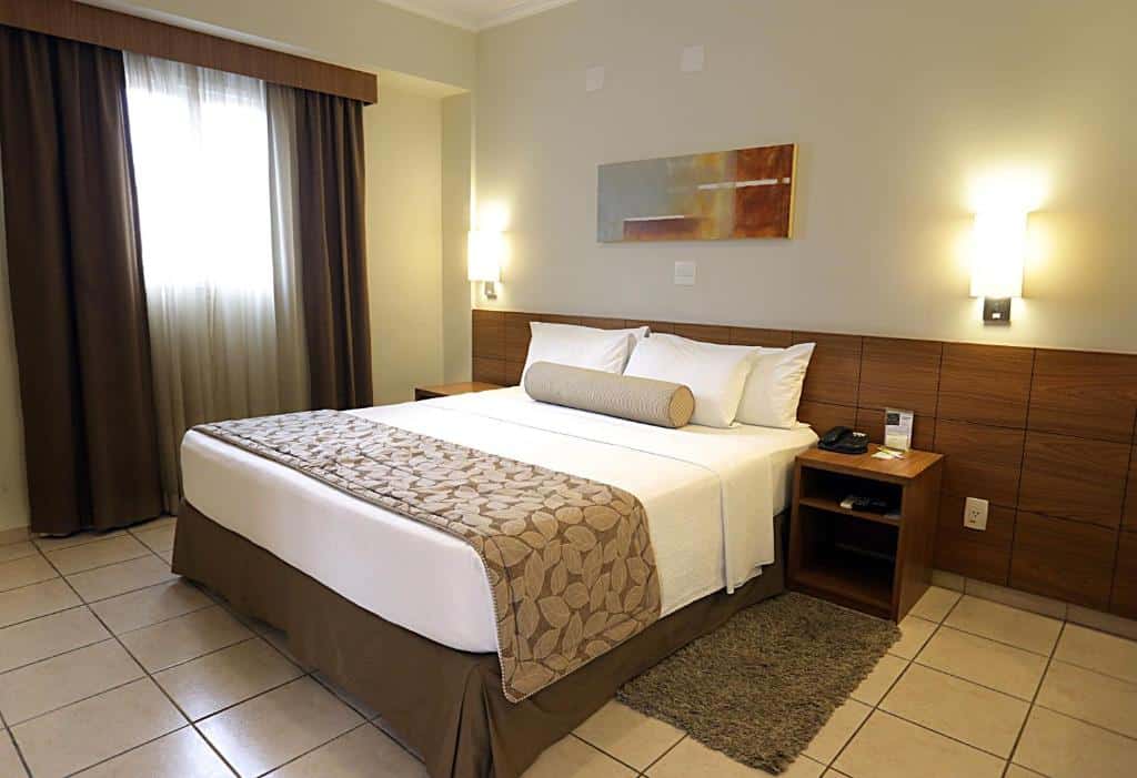 Quarto do Quality Hotel Jundiaí com uma cama de casal, duas mesinhas de cabeceira com luminárias, um pequeno tapete e uma janela com cortinas