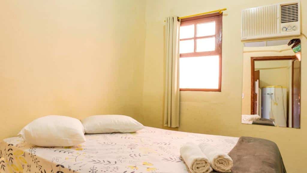 Quarto da Pousada Recanto Caiçara com cama de casal, janela de madeira com cortina bege e espelho no lado esquerdo.