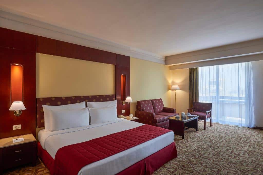 Quarto do Safir Hotel com cama de casal com cobertas brancas e vermelhas, duas cômodas com luminárias ao lado da cama, do lado esquerdo um sofá de dois lugares vermelho e uma poltrona vermelha com mesinha ao centro.
