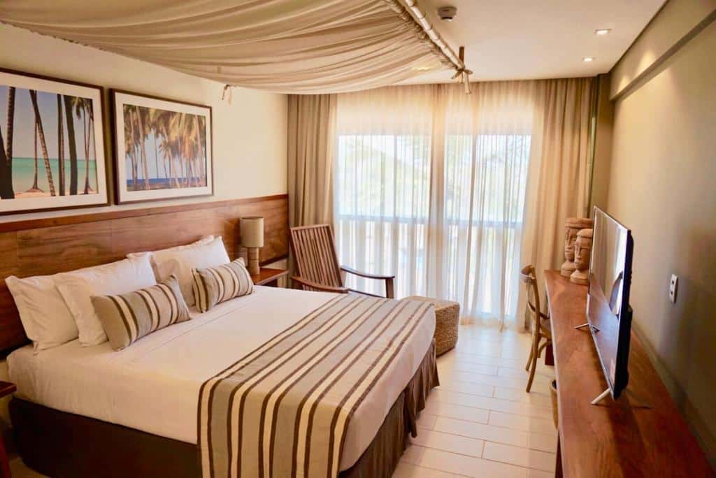 Quarto do Samoa Beach Resort com uma cama de casal, uma varanda com cortinas, um móvel de madeira com uma televisão, além e uma poltrona com lugar para apoiar os pés