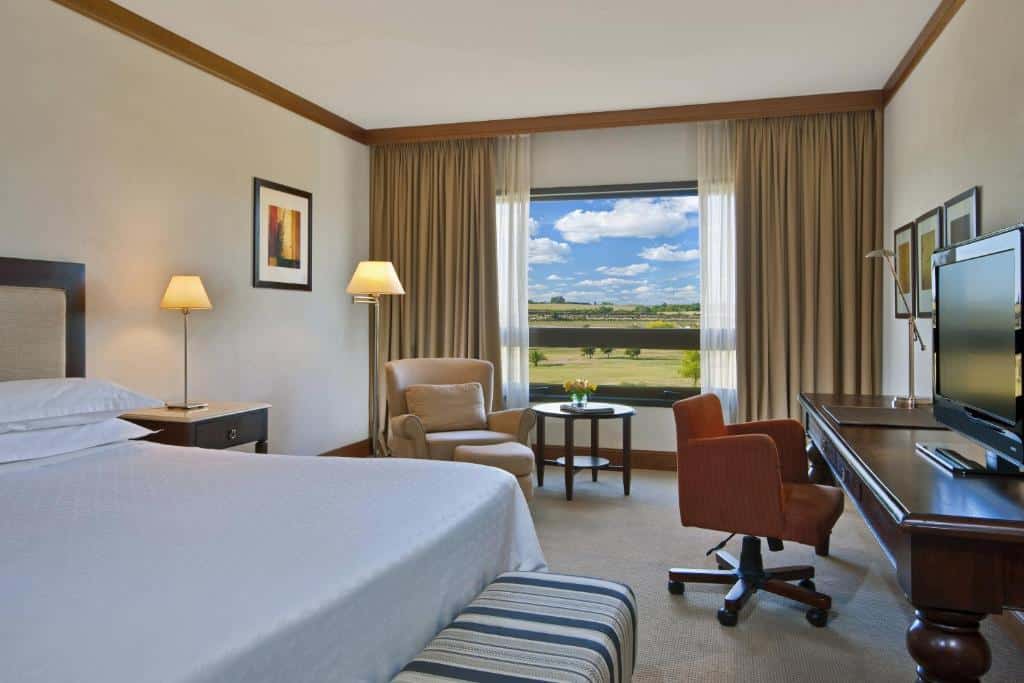 Quarto do Sheraton Colonia Golf & Spa Resort com uma janela ampla dando vista para as montanhas, do lado interno há uma cama de casal, uma poltrona, uma mesa de trabalho com uma televisão e luminária