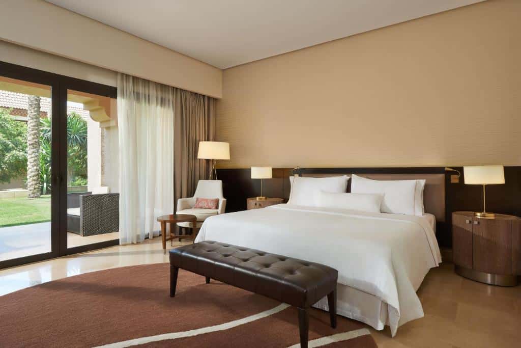 Quarto do The Westin Golf Resort & Spa com cama de casal do lado direito com duas cômodas de madeira com luminária, poltrona branca com mesa redonda de centro do lado esquerdo do quarto.