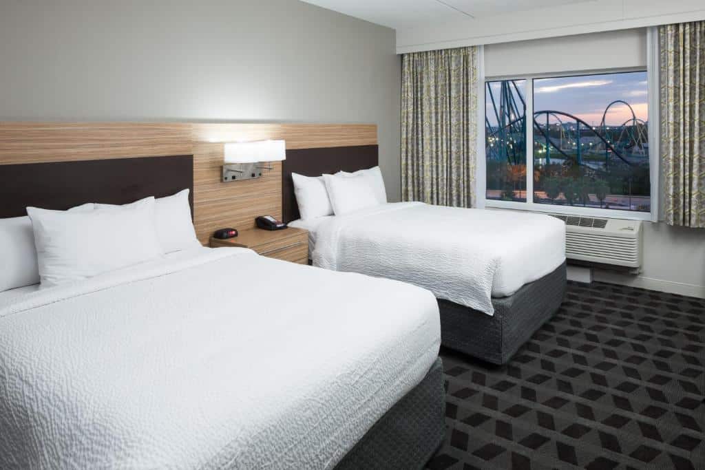 Quarto do TownePlace Suites by Marriott Orlando at SeaWorld com duas camas, uma de casal e outra de solteiro, o chão é de carpete e há uma janela com vista para o parque, e um ar-condicionado logo abaixo da janela