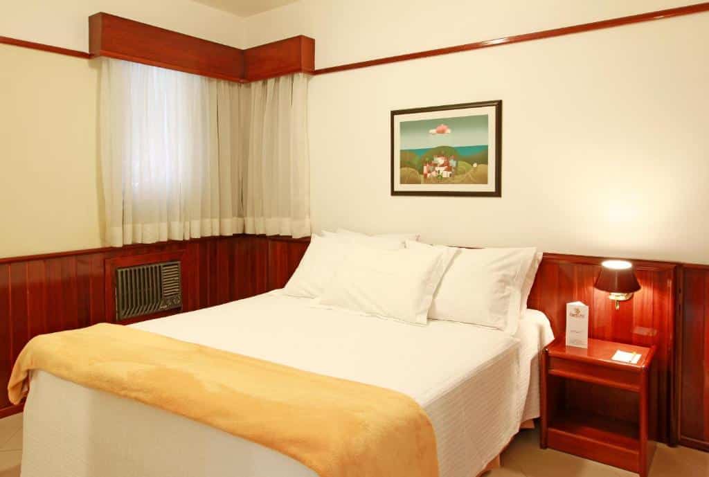 Quarto do Travel Inn Saint Charles com uma cama de casal, uma janela com cortinas, uma mesinha de cabeceira com abajur e um ar-condicionado, para representar hotéis perto do Hopi Hari