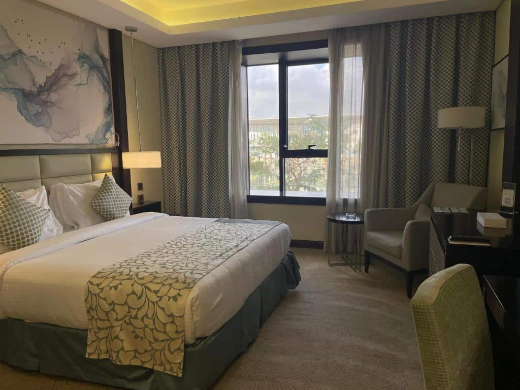 Quarto do Triumph Luxury Hotel com cama de casal, janelas com cortinas cinzas e brancas do lado esquerdo, poltrona bege do lado esquerdo e uma cômoda de madeira em frente a cama.