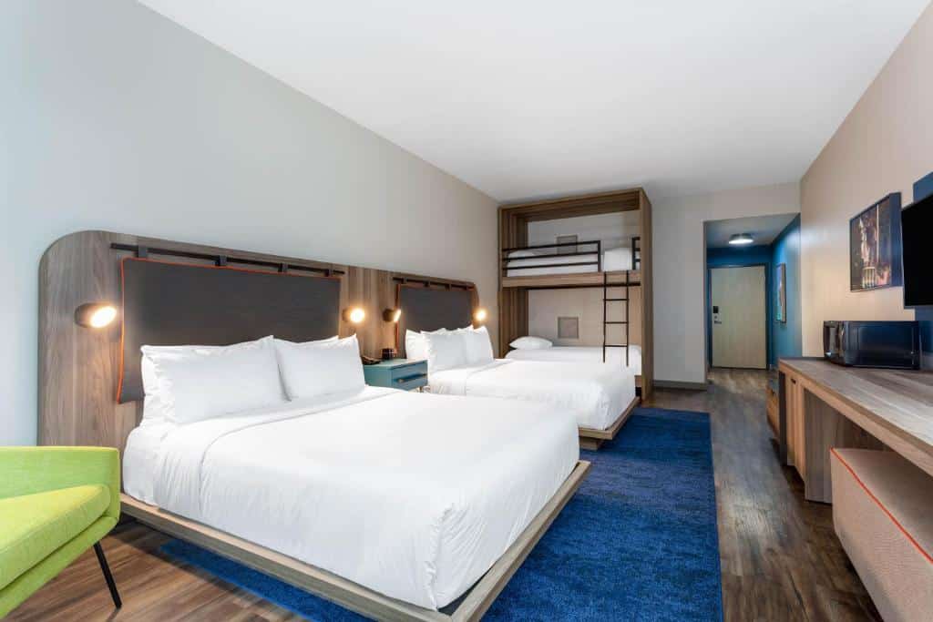 Quarto do TRYP by Wyndham Orlando com duas camas de casal e uma beliche, o chão é de madeira e há um amplo tapete azul marinho, além de um móvel de madeira com televisão, na cabeceira das camas há algumas luminárias presas