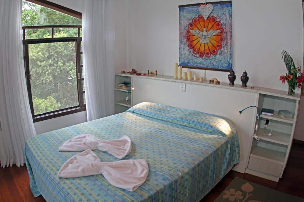Quarto do Venere - Bed and Breakfast com uma cama de casal, uma cabeceira com estantes, uma janela ampla com cortinas brancas e alguns itens de decoração pelo ambiente, para representar pousadas na Praia do Campeche