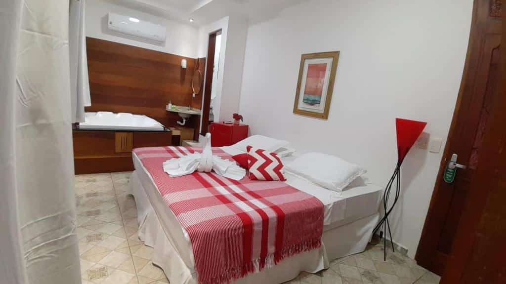 Quarto da Pousada Vila Barequeçaba com cama de casal, luminária do lado direito, do lado esquerdo frigobar vermelho e ao fundo banheira de hidromassagem.