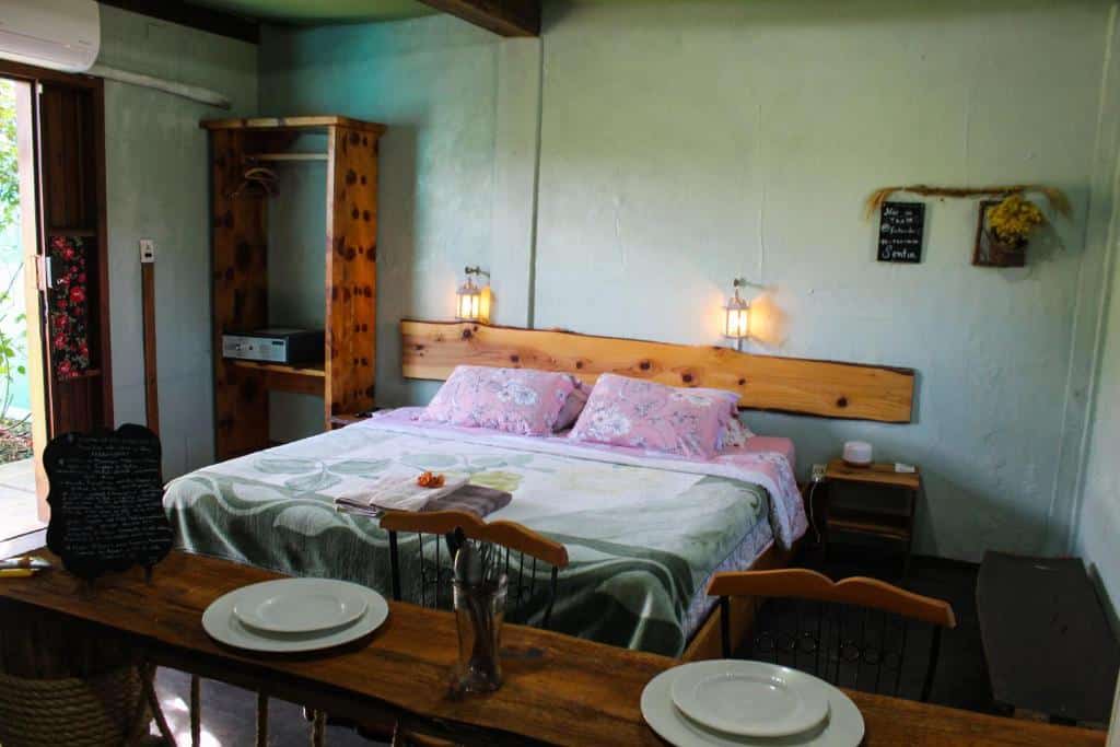 quarto da Vila Ecológica Pousada com uma cama de casal, uma mesa estreita de madeira rústica, duas cadeiras altas e uma porta com vista para a varanda à esquerda