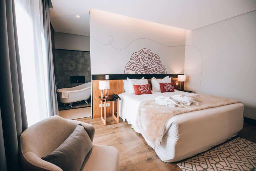 Quarto na Villa Amistà Campos do Jordão, com uma banheira, uma janela com cortinas e uma poltrona, há uma cama de casal, o chão é de madeira e há algumas almofadas em cima da cama