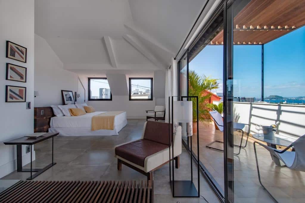 Quarto muito amplo na Villa Paranaguá Hotel & Spa com duas janelas e uma sacada grande com vista para a baía e a cidade, no ambiente há uma cama de casal, duas poltronas, abajures e quadros, tudo em estilo minimalista