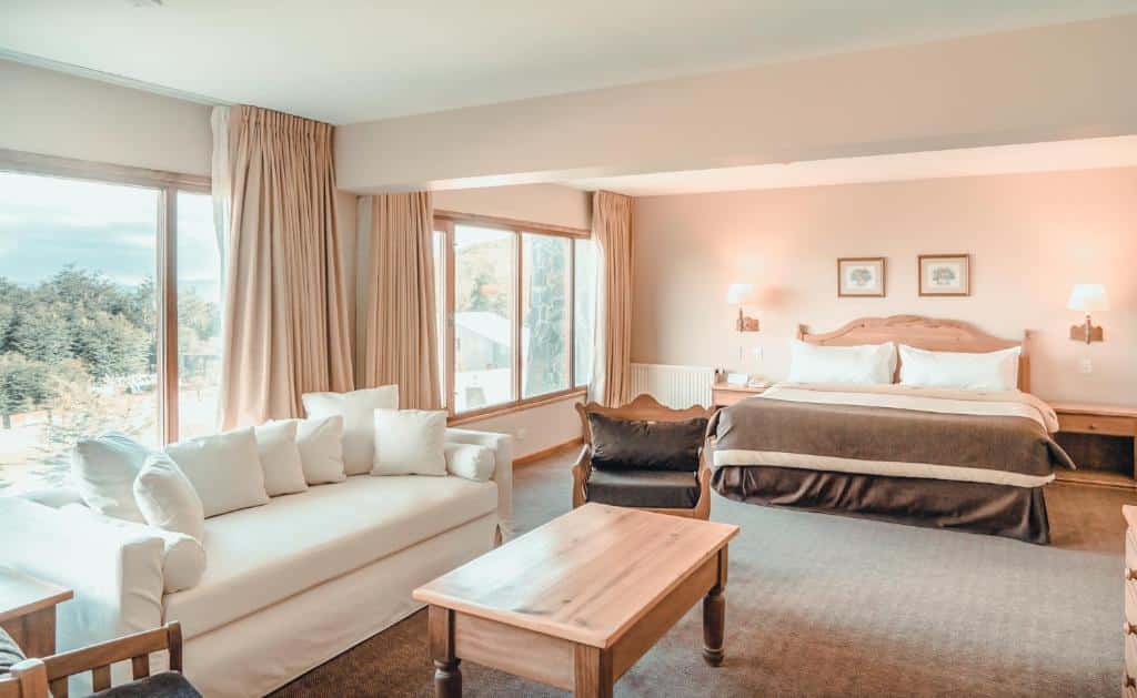 suíte do Wyndham Garden Ushuaia Hotel del Glaciar com uma sala de estar, janelas amplas com vista para a natureza no canto esquerdo e uma cama de casal no canto direito, ao fundo