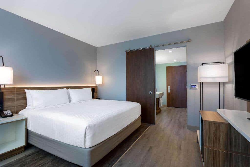 Quarto do Wyndham Garden Miami International Airport com cama de casal, duas cômodas ao lado da cama com luminária, em frente a cama TV na parede com uma cômoda de madeira abaixo.