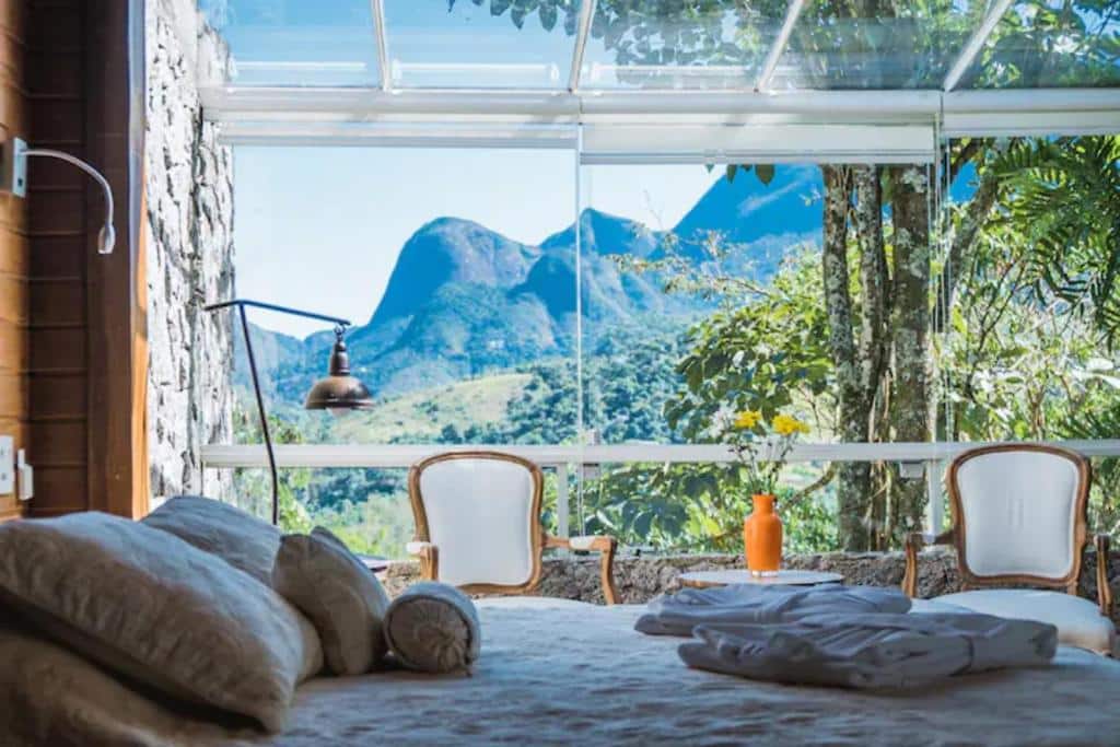 Janela ampla de vidro em um dos quartos da Quinta 59, é possível ver diversas montanhas e muito verde, dentro do quarto há uma cama de casal e duas poltronas de frente para essa vista, para representar chalés em Petrópolis