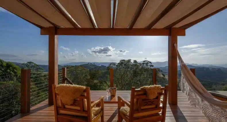 Uma varanda ampla de madeira na Quinta dos Pinhais Pouso e Charme com duas cadeiras de madeira e uma rede de descanso de frente para as montanhas, para representar pousadas para casal perto de SP