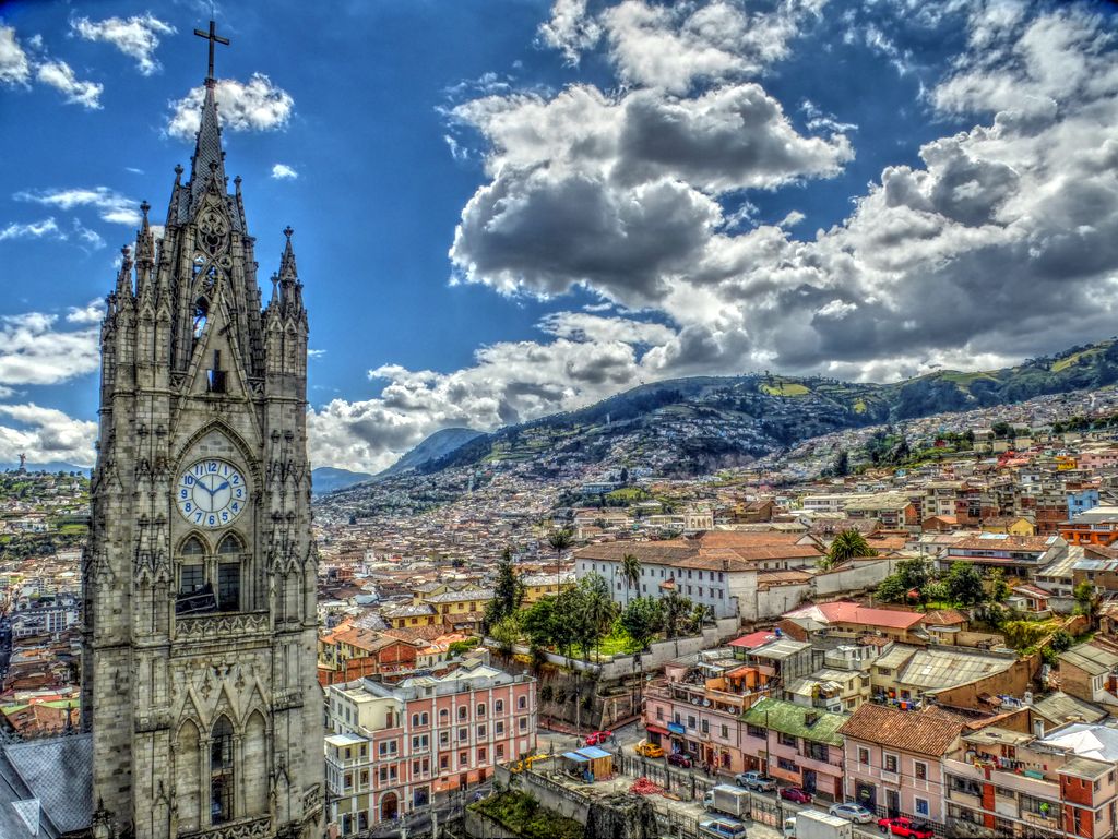 Vista do alto de uma região de Quito, com várias casas e a torre de uma igreja medieval, com um relógio redondo. Há montanhas ao fundo e o céu está com algumas nuvens em um dia ensolarado. Imagem para ilustrar o post de chip celular Quito