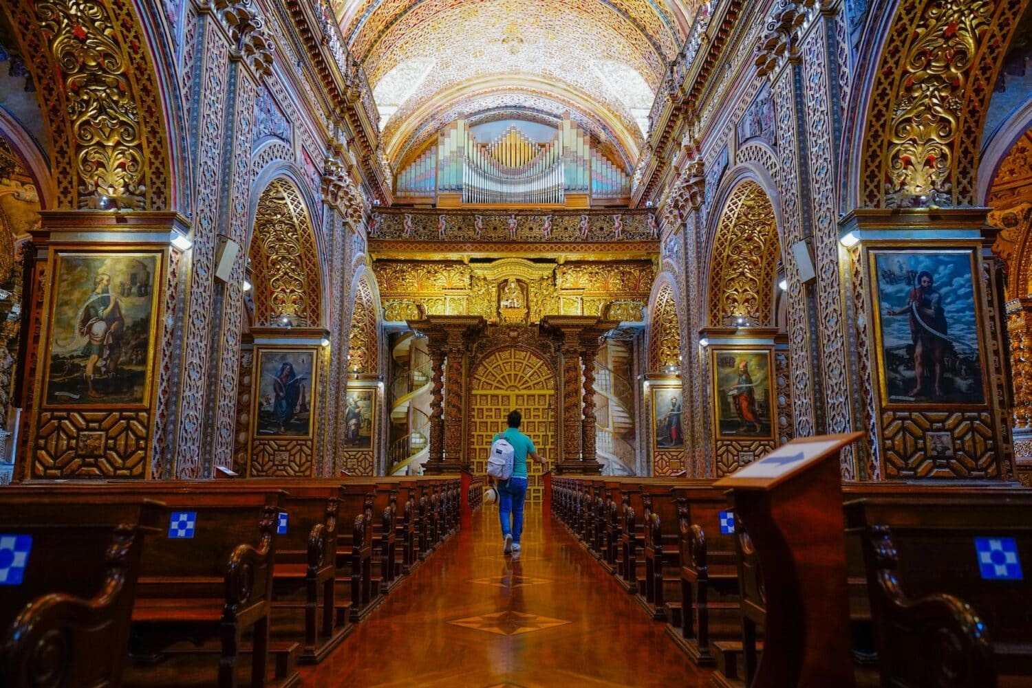 Homem vestido de blusa azul e calça jeans com mochila branca nas costas e segurando um chapéu em uma das mãos, andando de costas no corredor de uma igreja cheia de obras de arte e teto todo enfeitado em Quito, Equador. Imagem para ilustrar o post de chip de celular Quito