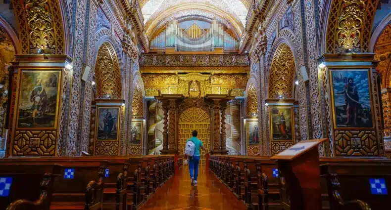 Homem vestido de blusa azul e calça jeans com mochila branca nas costas e segurando um chapéu em uma das mãos, andando de costas no corredor de uma igreja cheia de obras de arte e teto todo enfeitado em Quito, Equador. Imagem para ilustrar o post de chip de celular Quito