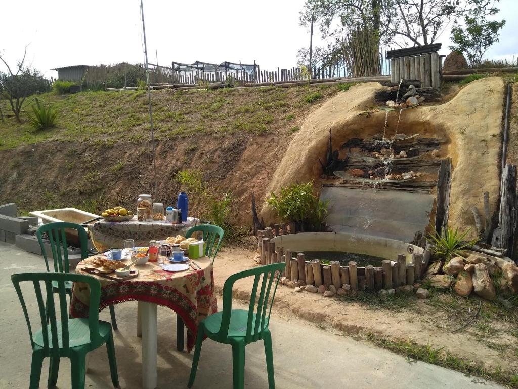 Cascata rústica do Recanto Cheio de Cheiro, uma das pousadas em Guararema, com bastante natureza em volta e duas mesas à frente com cadeiras verdes e itens de café da manhã em cima.