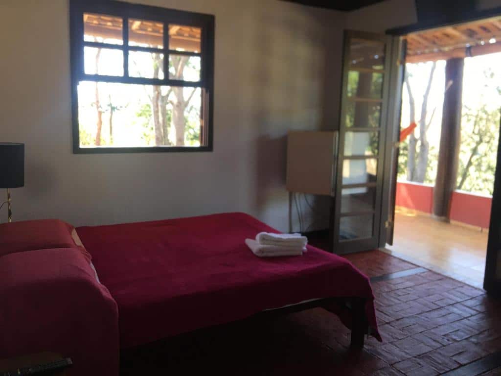 Quarto da Recanto das Acácias, com uma cama de casal com lençol vermelho em cima, janela do lado esquerdo e porta aberta dando acesso à varanda com vista para natureza