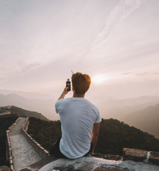 Homem de costas sentado no topo da Muralha da China. Ele segura um celular enquanto tira fotos da paisagem. - Foto: Road Trip with Raj via Unsplash