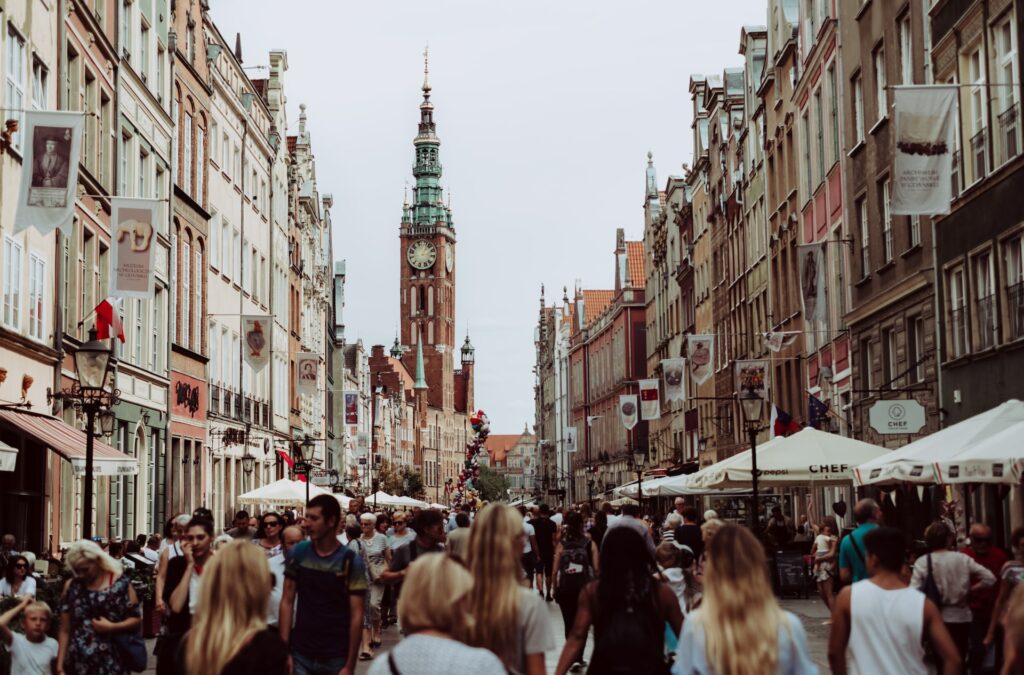 Pessoas andando em rua da cidade de Gdansk, na Polônia, um dos países integrantes do Tratado de Schengen. Em cada lado da rua há prédios baixos, de dois ou três andares, além de bandeiras na maioria deles. Ao fundo há uma torre, com relógio.