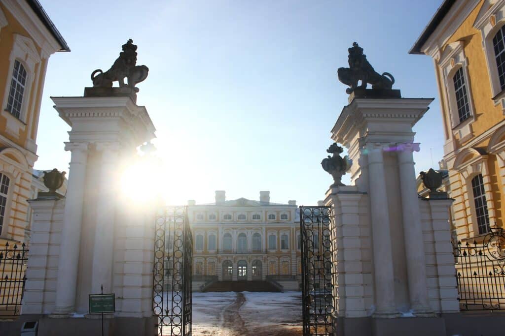 Portão de entrada para o palácio Rundāles Pils em tons de amarelo e branco, com sol durante o dia.