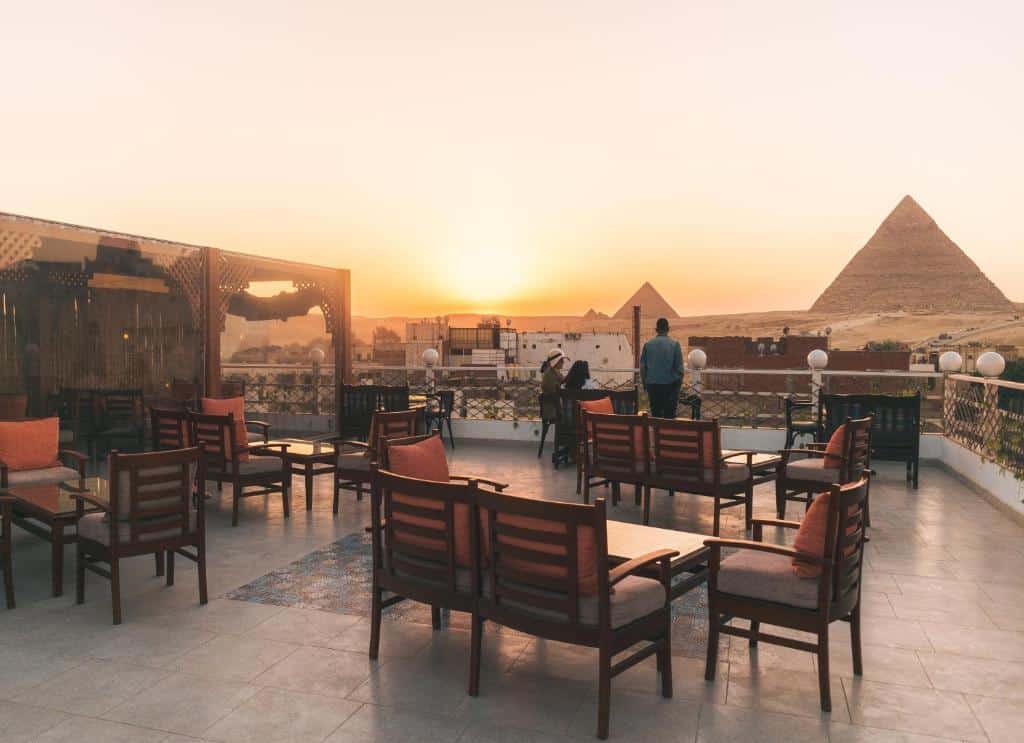 Área de lazer na sacada do Jimmy Pyramids View com cadeiras de madeiras com estofados vermelhos com vista para as pirâmides. Representa Hotéis no Cairo.