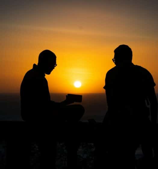 Silhueta preta de 4 pessoas em uma mureta olhando um pôr do sol alaranjado. Há dois homens usando óculos, sendo um com um celular na mão e outro olhando para esse celular. Imagem para ilustrar o post de chip celular Nicarágua