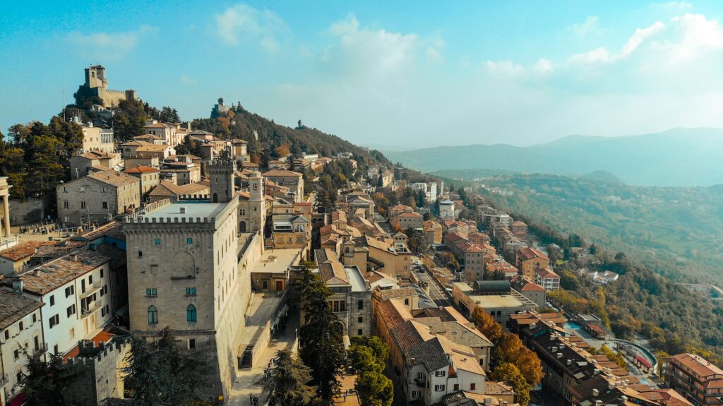 Vista de San Marino em uma região alta, que tem casas e construções históricas em uma montanha com vegetação em volta e bastante natureza nas redondezas. Está de dia e o céu ensolarado com poucas nuvens
