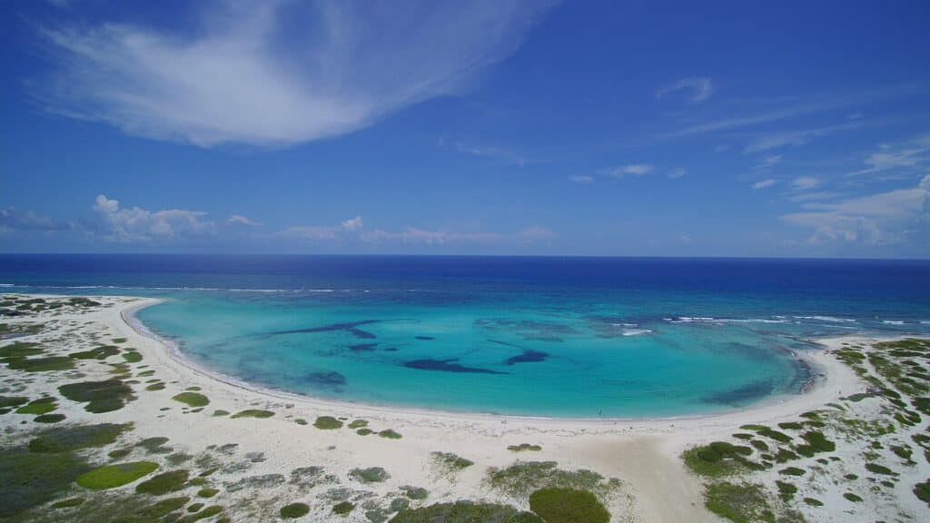 Vista panorâmica de praia paradisíaca em Aruba, na região de San Nicolas North. Há areia clara na orla, com chumaços de grama espalhados, e uma parte da água é mais clara e forma uma piscina, sendo que o mar atrás é mais escuro